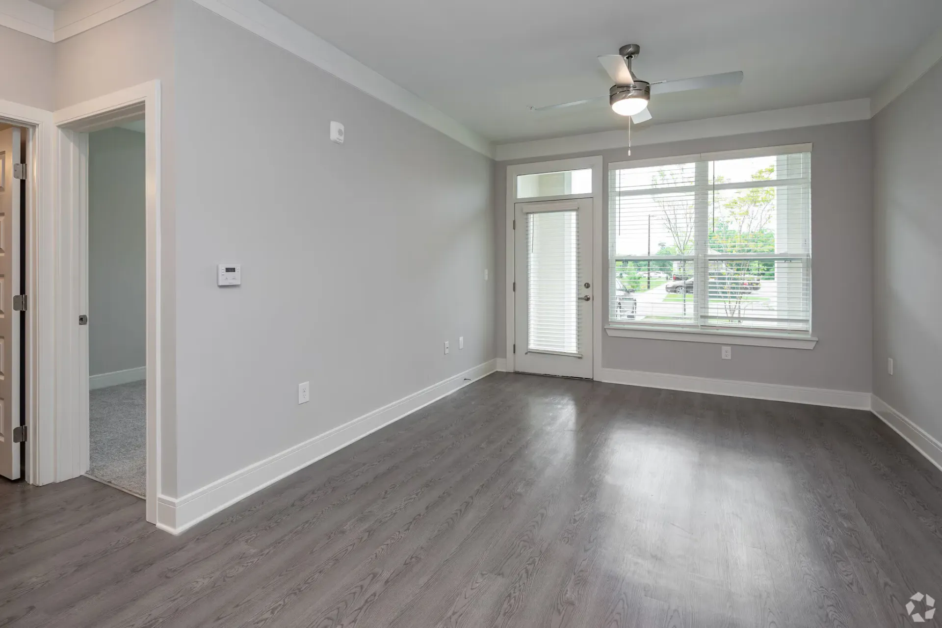 Empty living room with gray walls, large window, ceiling fan, and outside door.