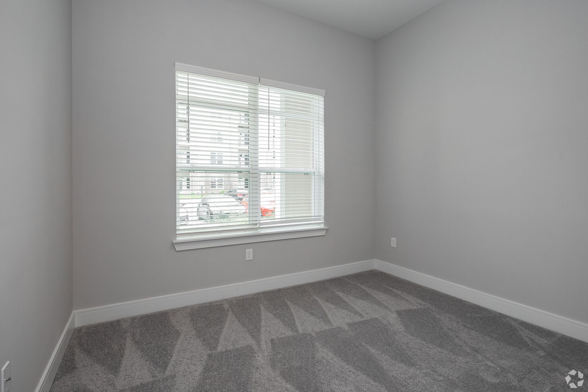 Empty bedroom with light gray walls, a window with blinds, and a carpeted floor.