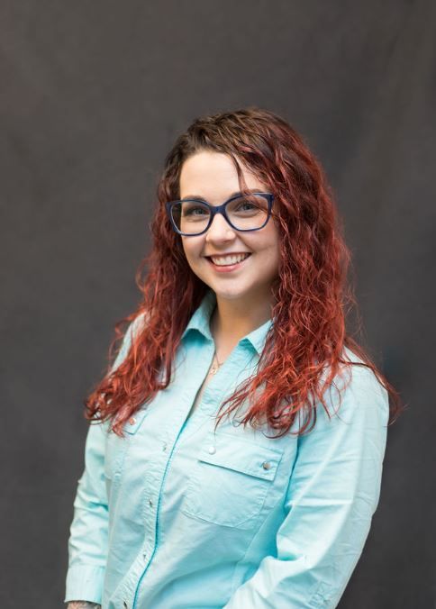 A woman with red hair and glasses is wearing a blue shirt and smiling.