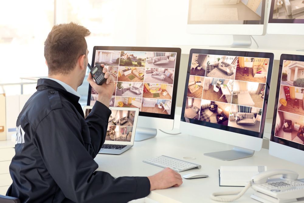 A security guard is sitting at a desk in front of three computer monitors.