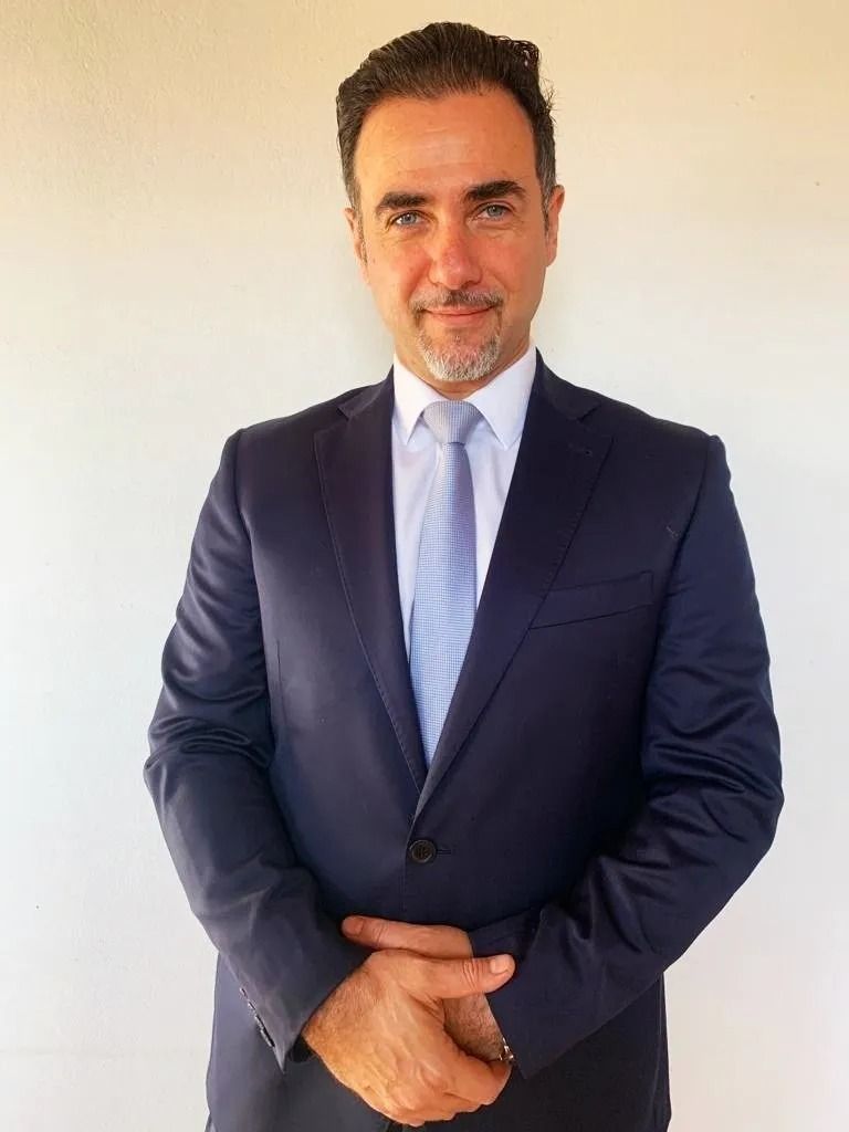 Un hombre con cabello corto y oscuro y barba, vestido con un traje azul marino