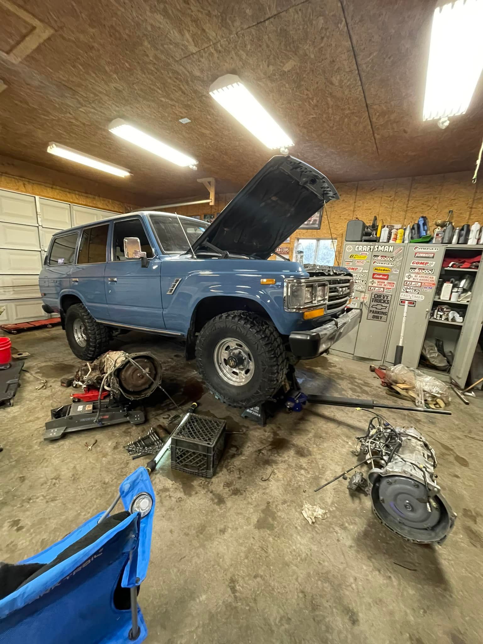 Blue Land Cruiser in a garage, hood up, surrounded by auto parts.