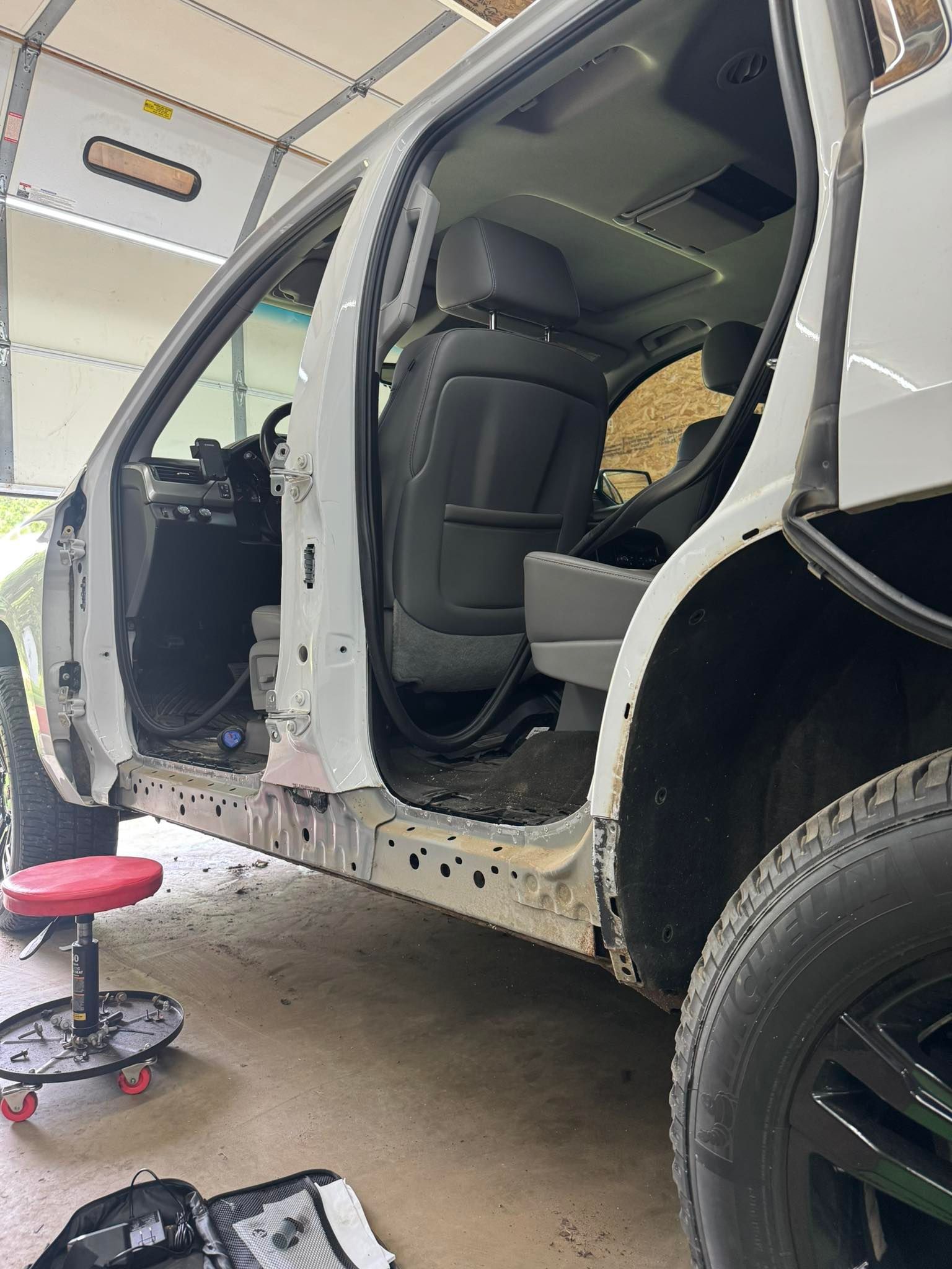 A white SUV undergoing repair, with open doors revealing interior and a red stool nearby.