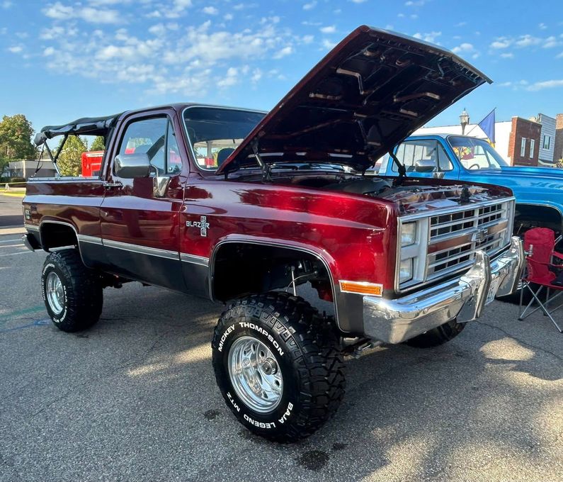 Burgundy 1980s Chevy Blazer with the hood up, parked at a car show, chrome wheels, and lifted suspension.