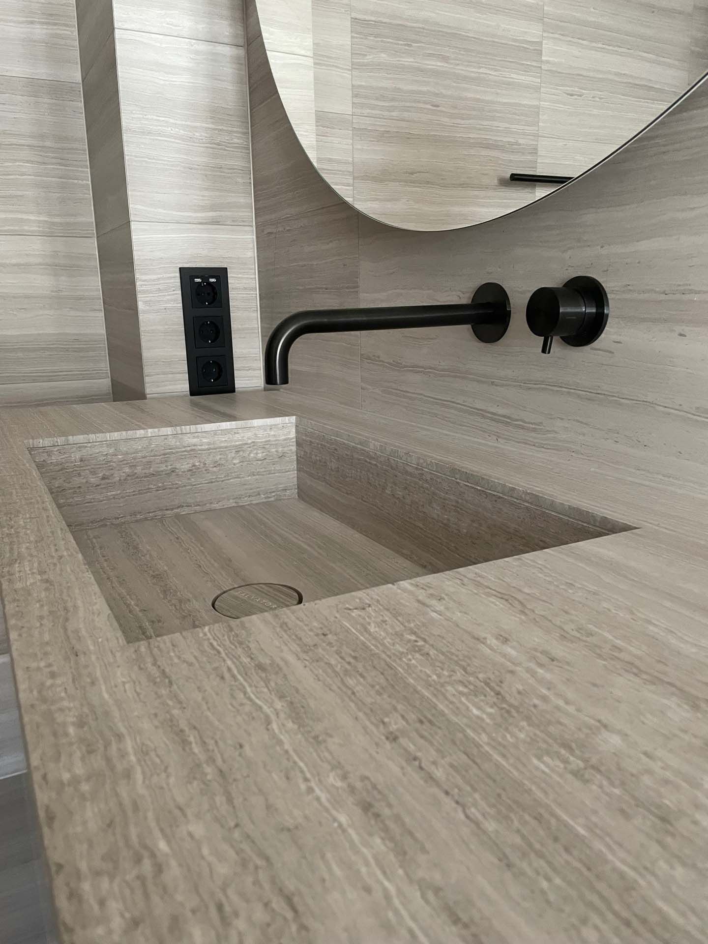 Ein Waschbecken aus Fliesen mit Holzoptik im Badezimmer mit einem schwarzen Wasserhahn und einem Spiegel.