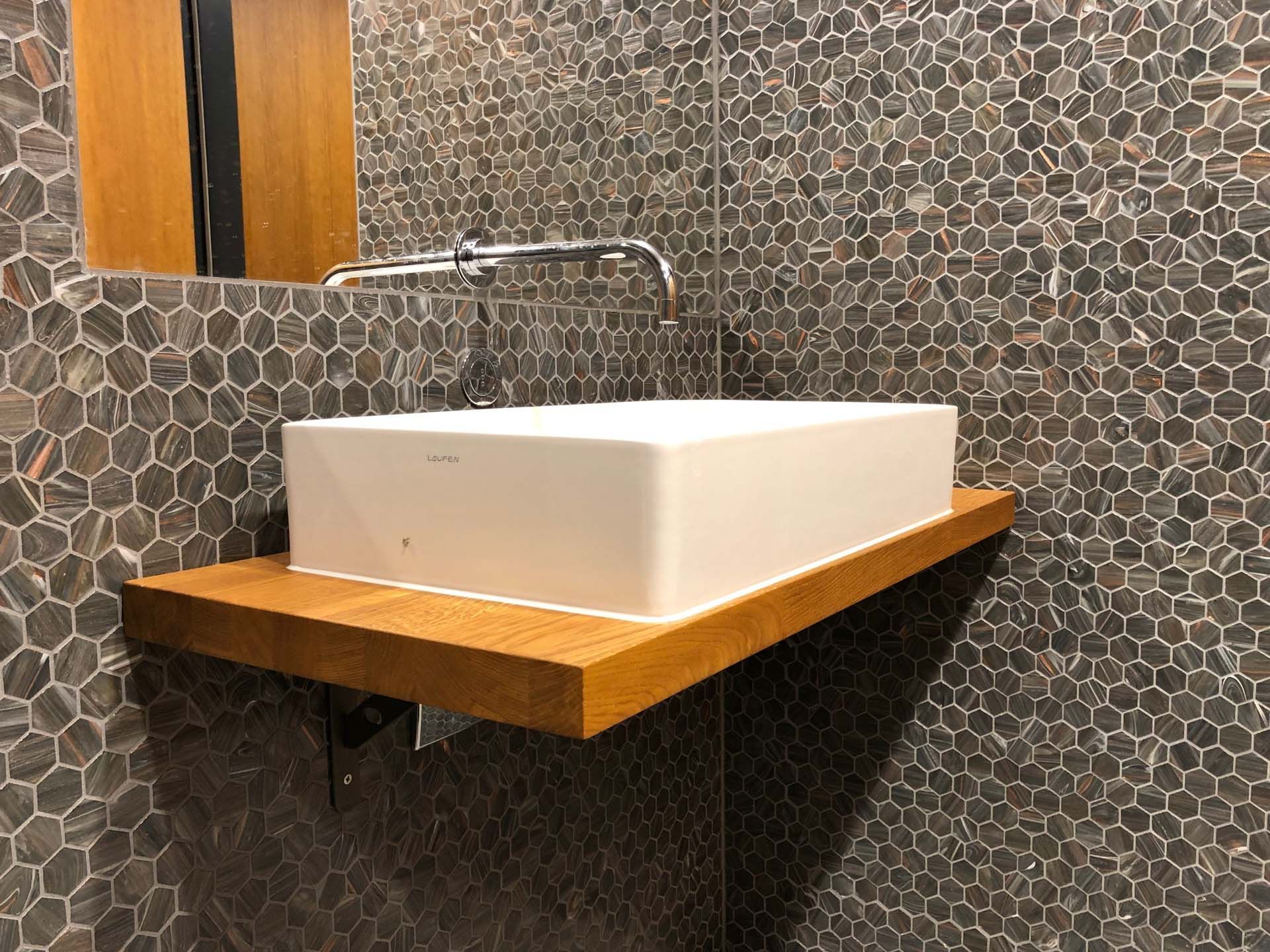 Ein Badezimmer, mit Fliesen im Mosaikmuster an den Wänden, mit einem Waschbecken und einem Spiegel auf einem Holzregal.