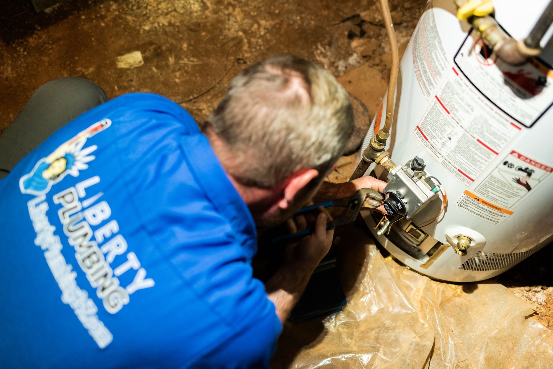 A man wearing a blue shirt that says liberty plumbing is working on a water heater.