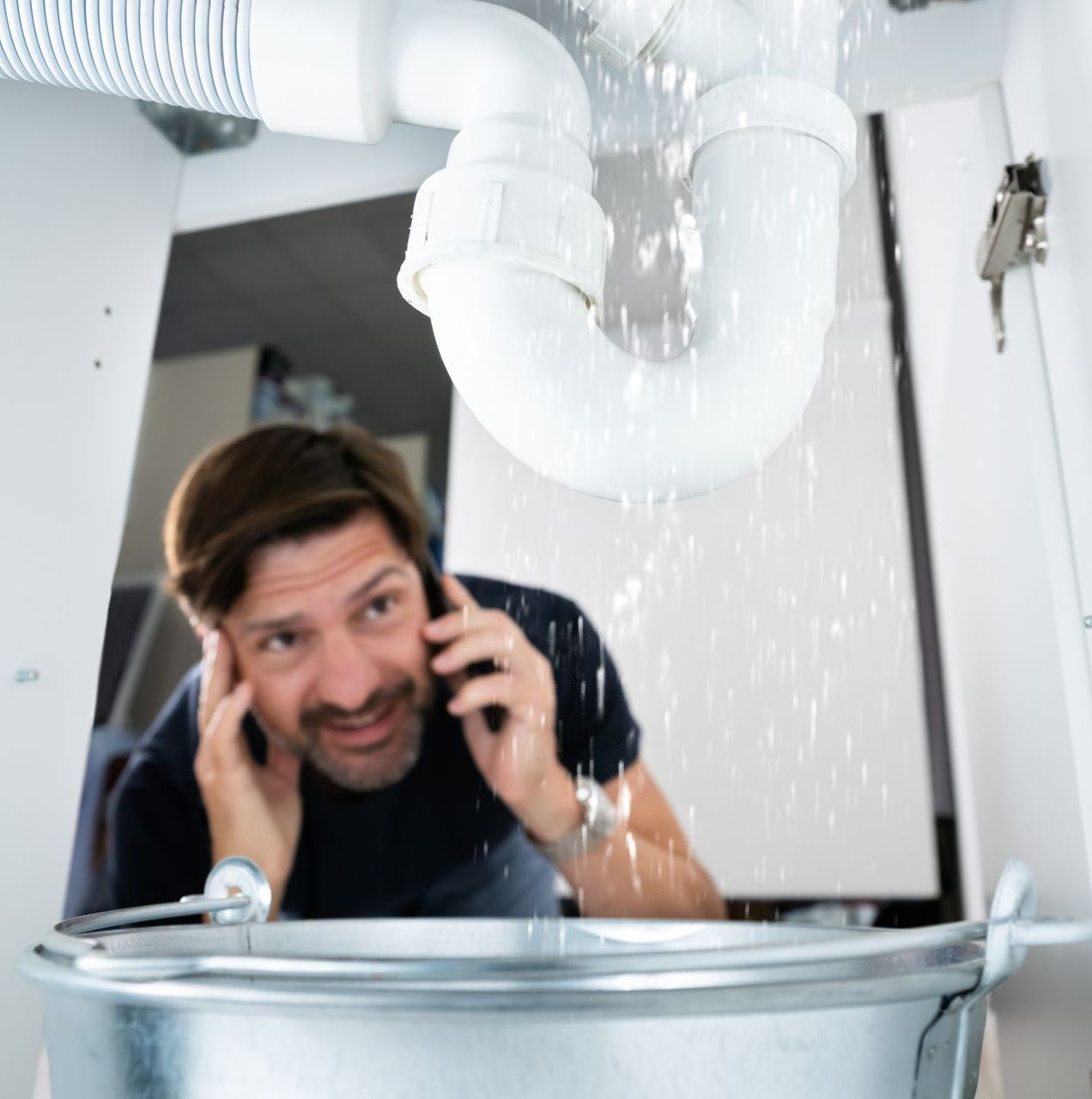 A man is talking on a cell phone under a sink with water coming out of it