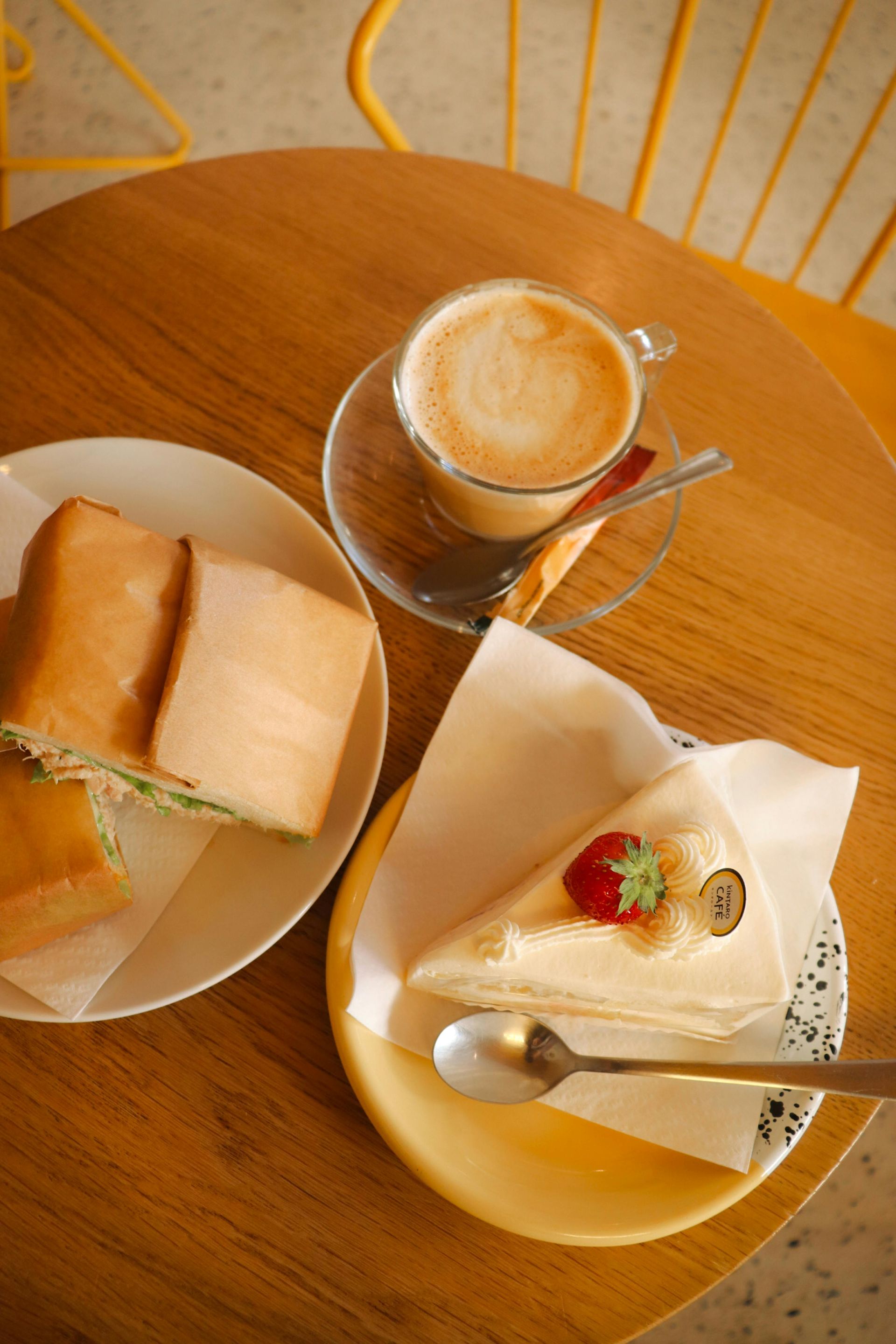 Coffee, cake, and sandwich on a wooden table. Yellow chair in background.
