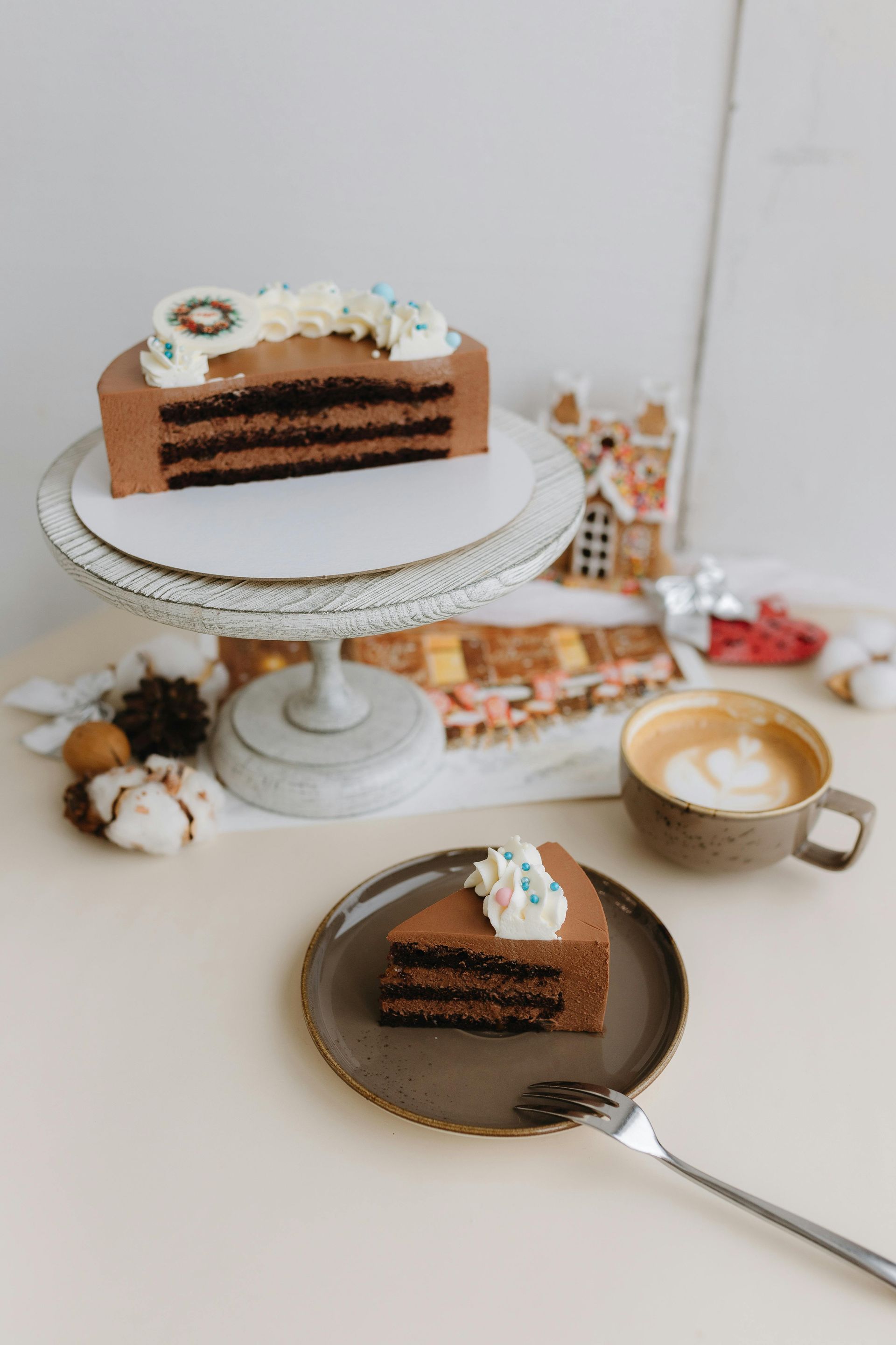 Chocolate cake slice on plate, cake on stand, latte, and gingerbread house on table.