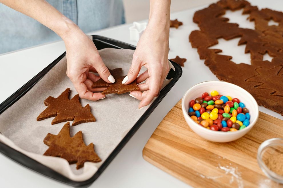 Woman placing tree-shaped cookies on a baking sheet, alongside bowl of colorful candies and cut-out dough.