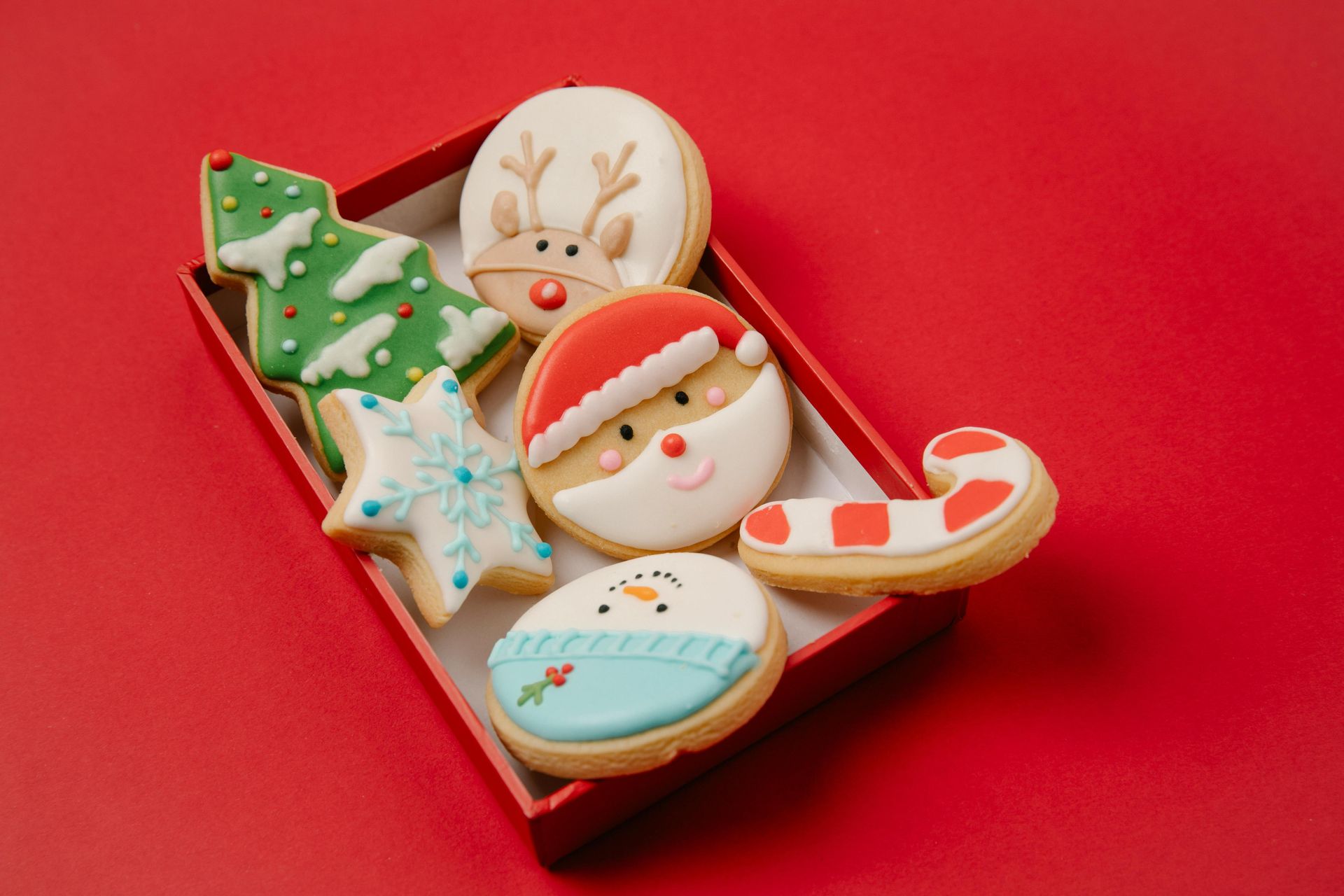 Christmas cookies in a red box: decorated tree, reindeer, Santa, snowflake, snowman, and candy cane on a red background.