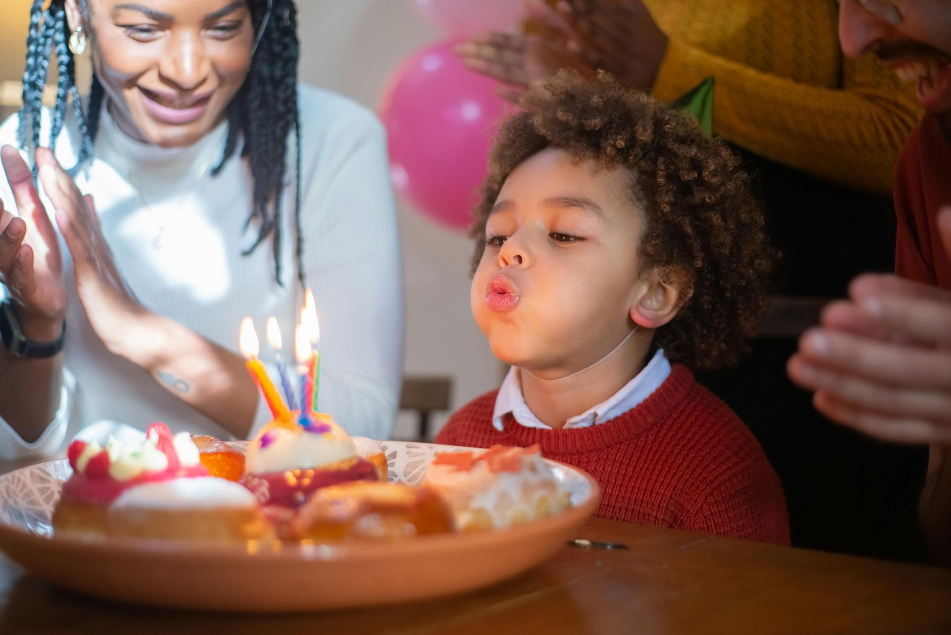 Young child blowing out birthday candles on donuts, people clapping, smiling.