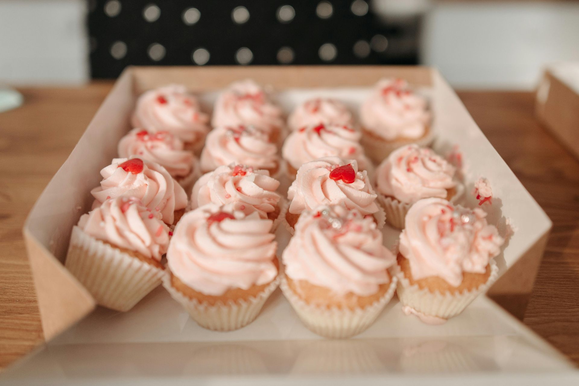Box of pink frosted cupcakes with red sprinkles, on a wooden surface.