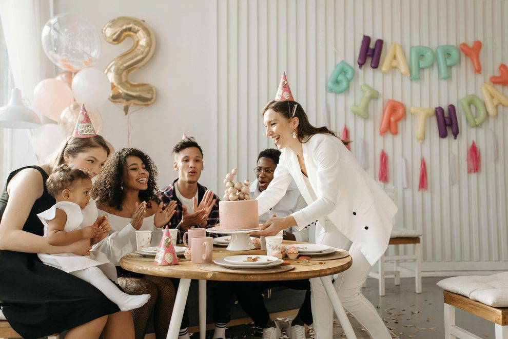 People at a birthday party; woman holding a cake. Balloons and 