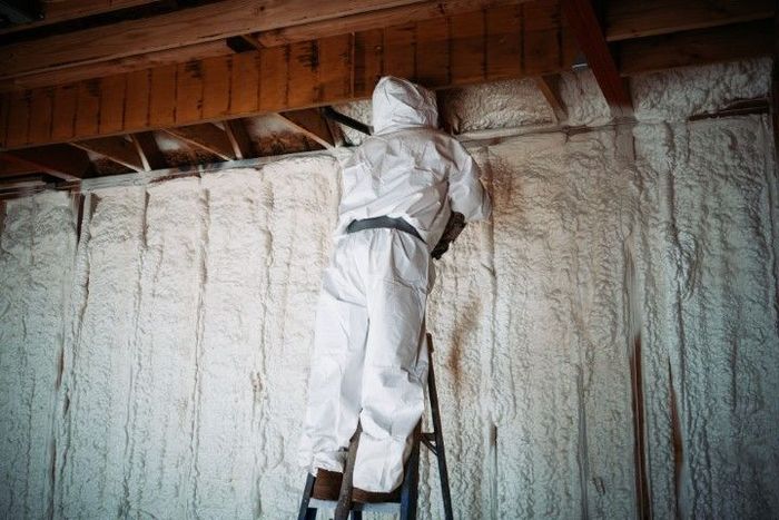 Person in protective suit spraying insulation foam on a wall, standing on a ladder.