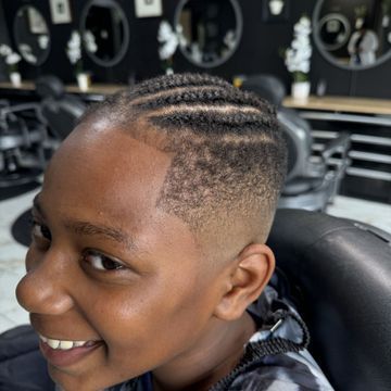 A young boy is sitting in a barber chair with braids in his hair.