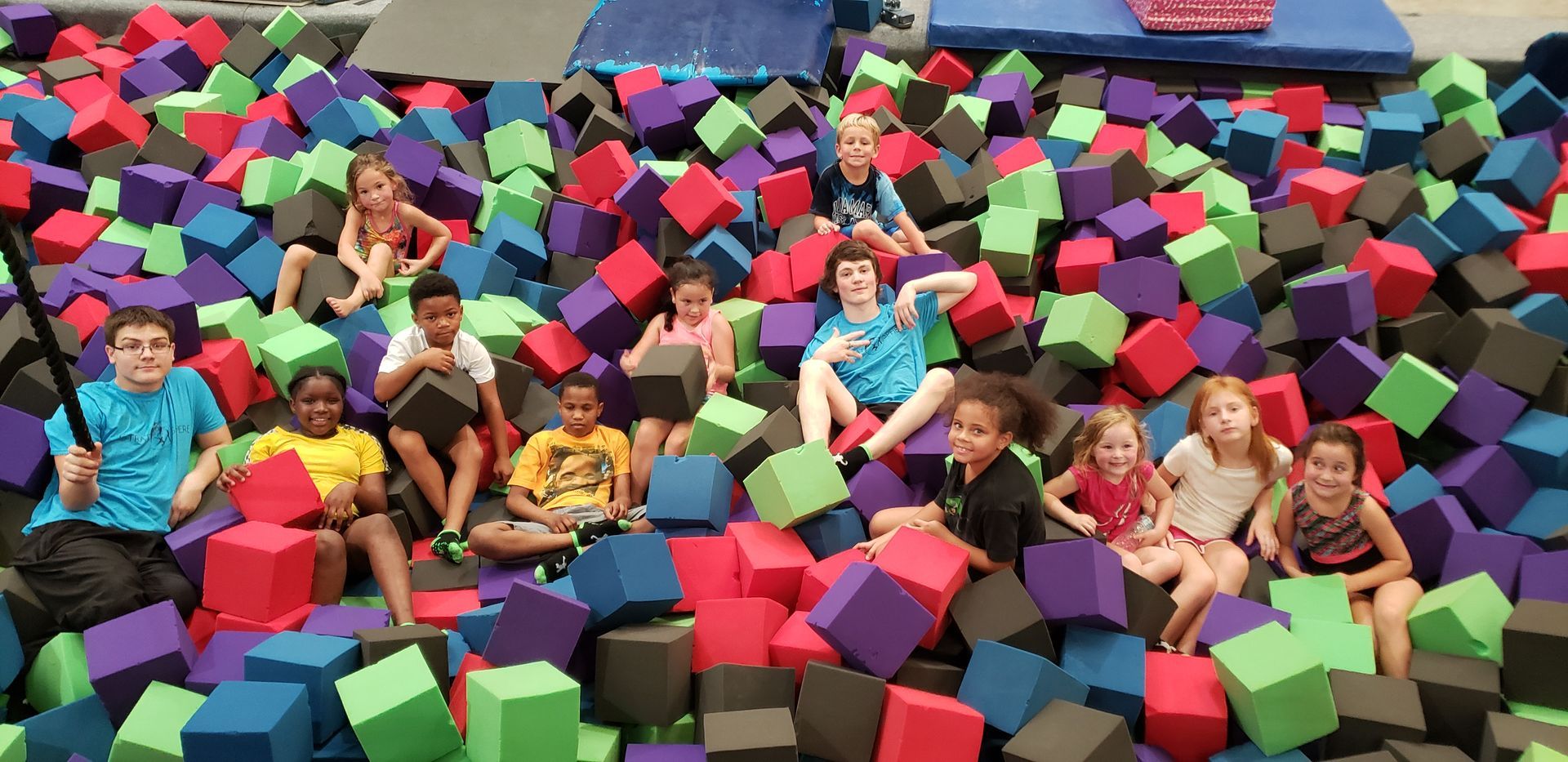 Children sitting in a pit of colorful foam cubes. Some hold blocks, and some smile.
