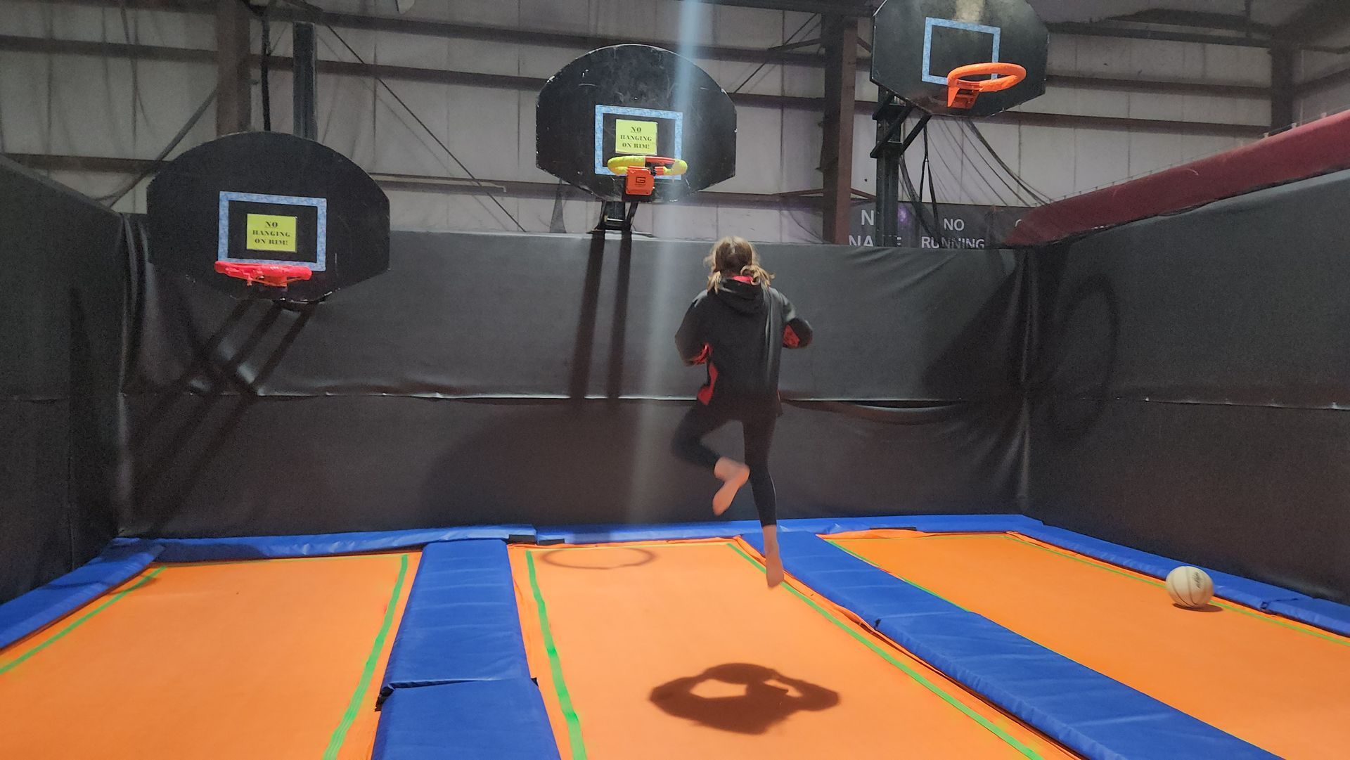 A person jumping on a trampoline towards a basketball hoop. Indoor trampoline park setting, with two hoops and a ball.