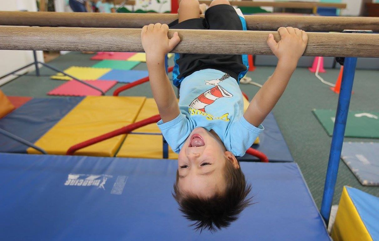 Child hanging upside down on a wooden bar in a gym, smiling.