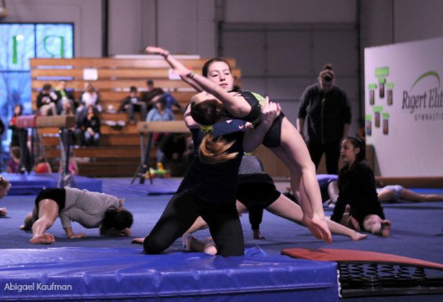 Gymnast being assisted with backbend by another gymnast on blue mat in a gym.