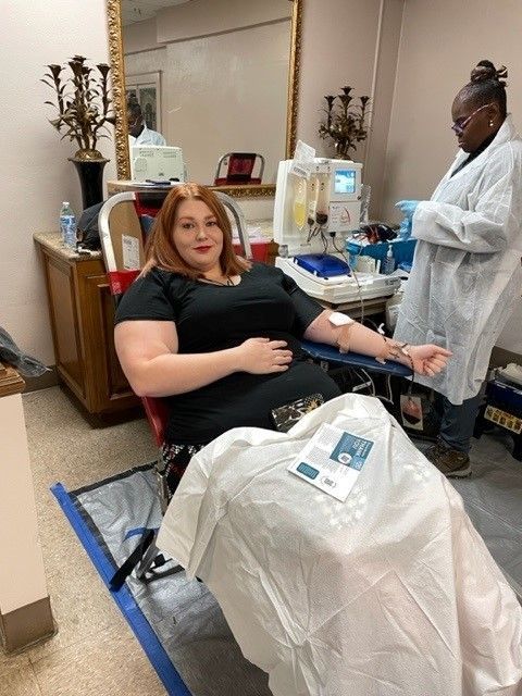 Person sitting while donating blood, with a nurse in the background.
