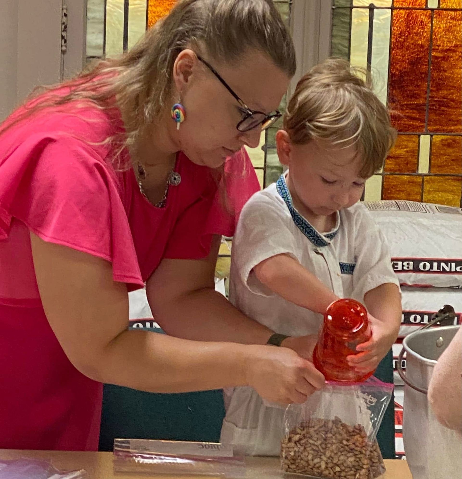 Woman in pink dress helps child fill a bag with trail mix indoors.