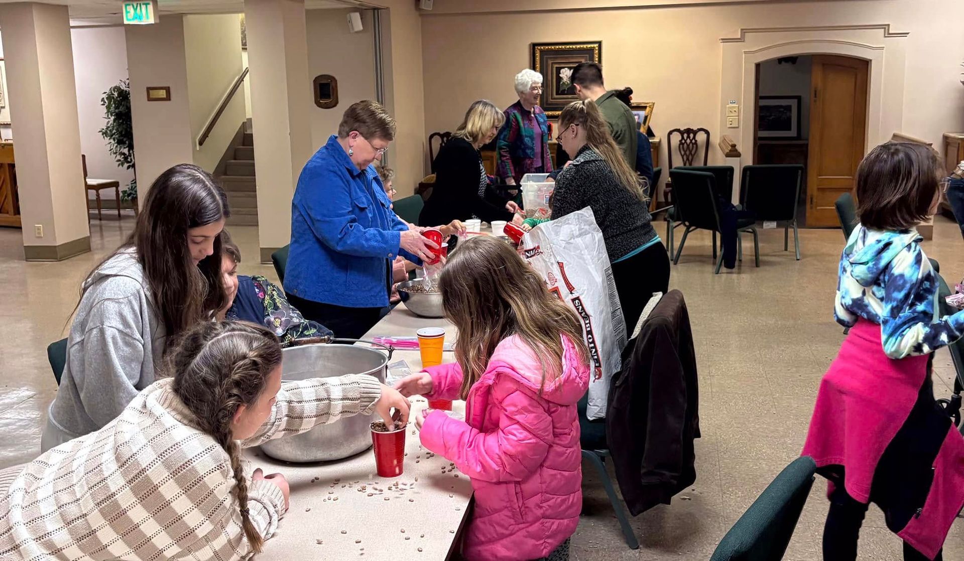 People gather around tables, likely at an event. A woman in blue pours from a container. Children and adults participate.
