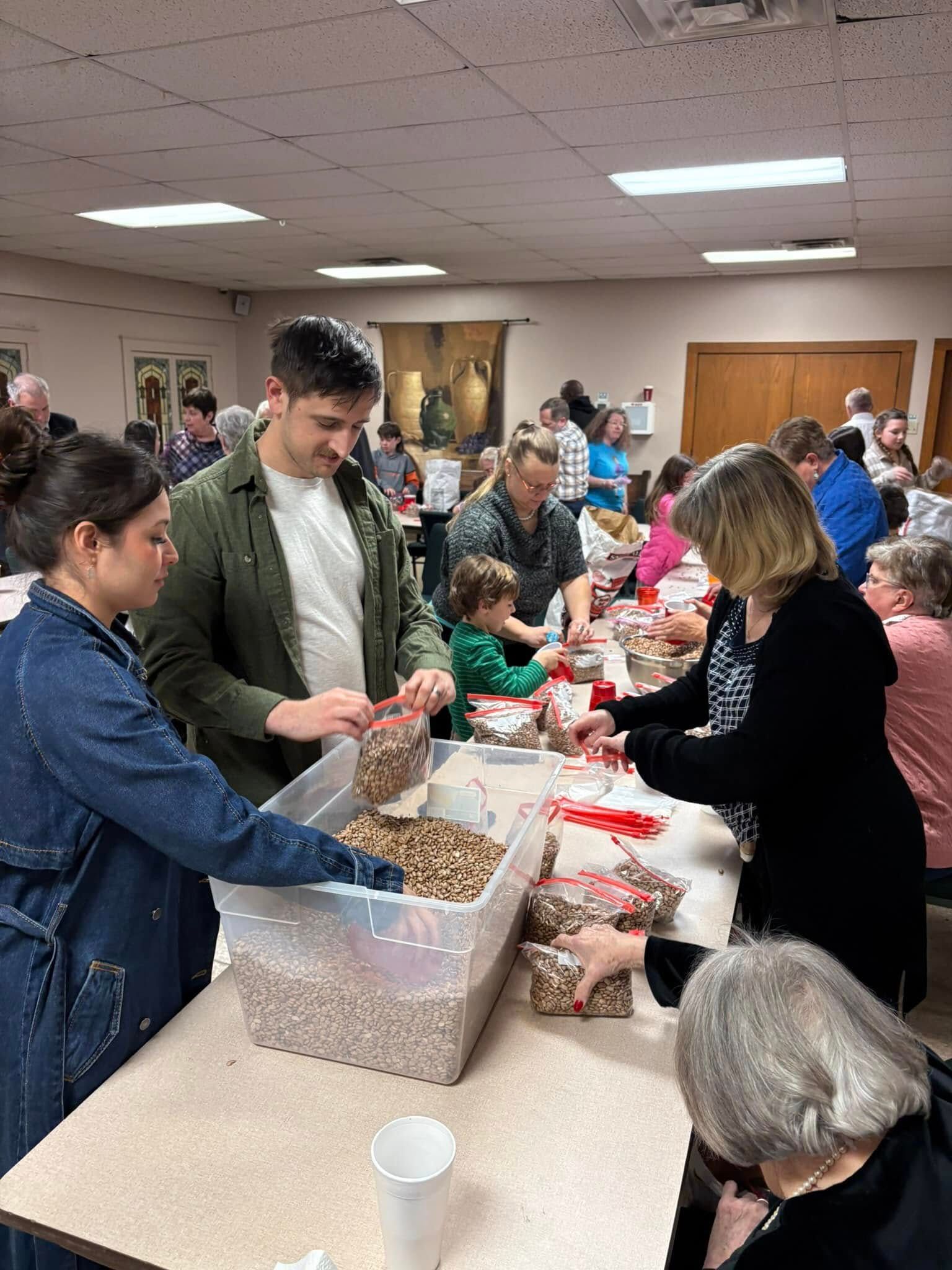 People filling bags with food at a community event. Indoor setting with tables, containers, and volunteers.