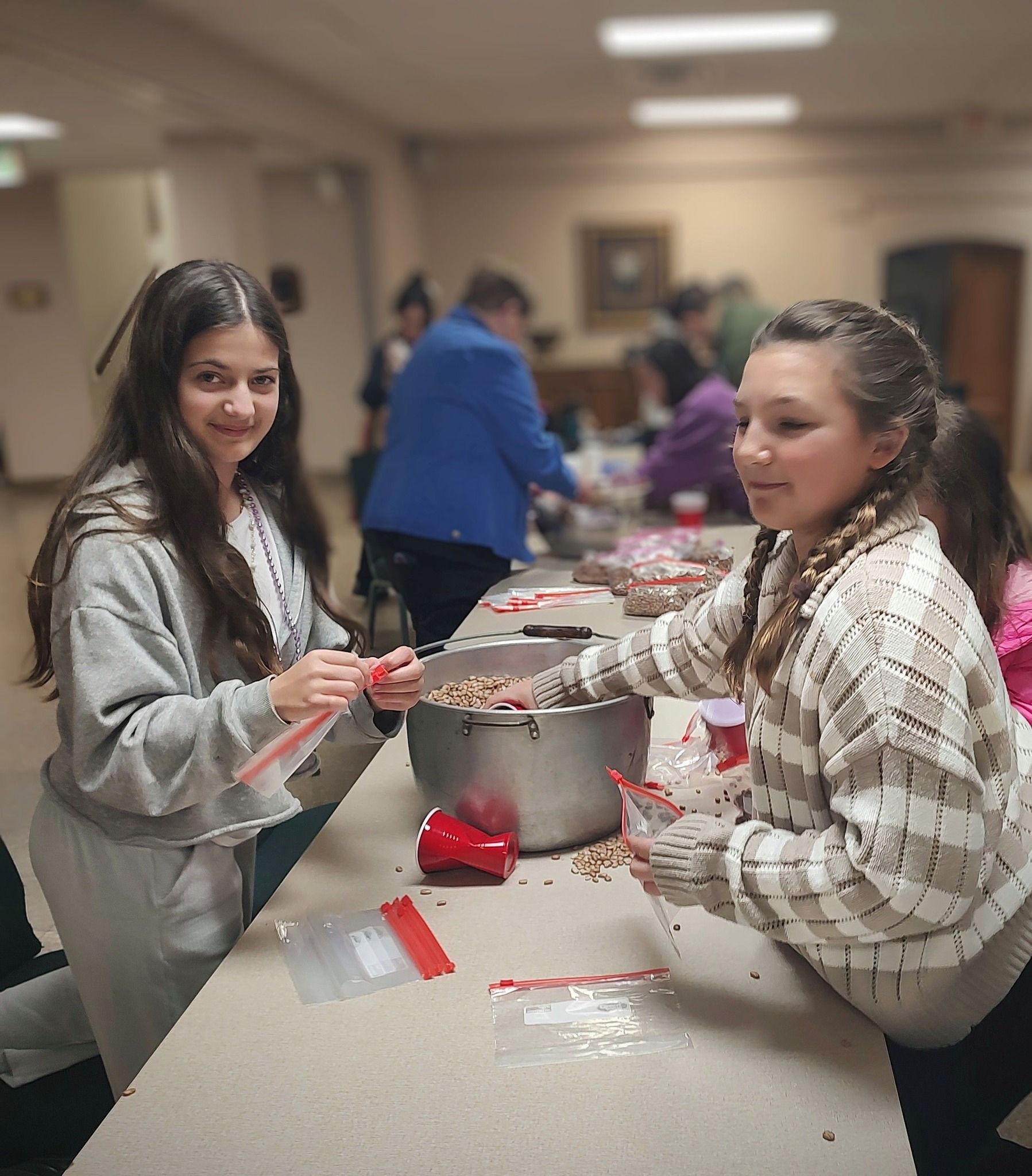 Two girls filling bags with food at a table, people in background.