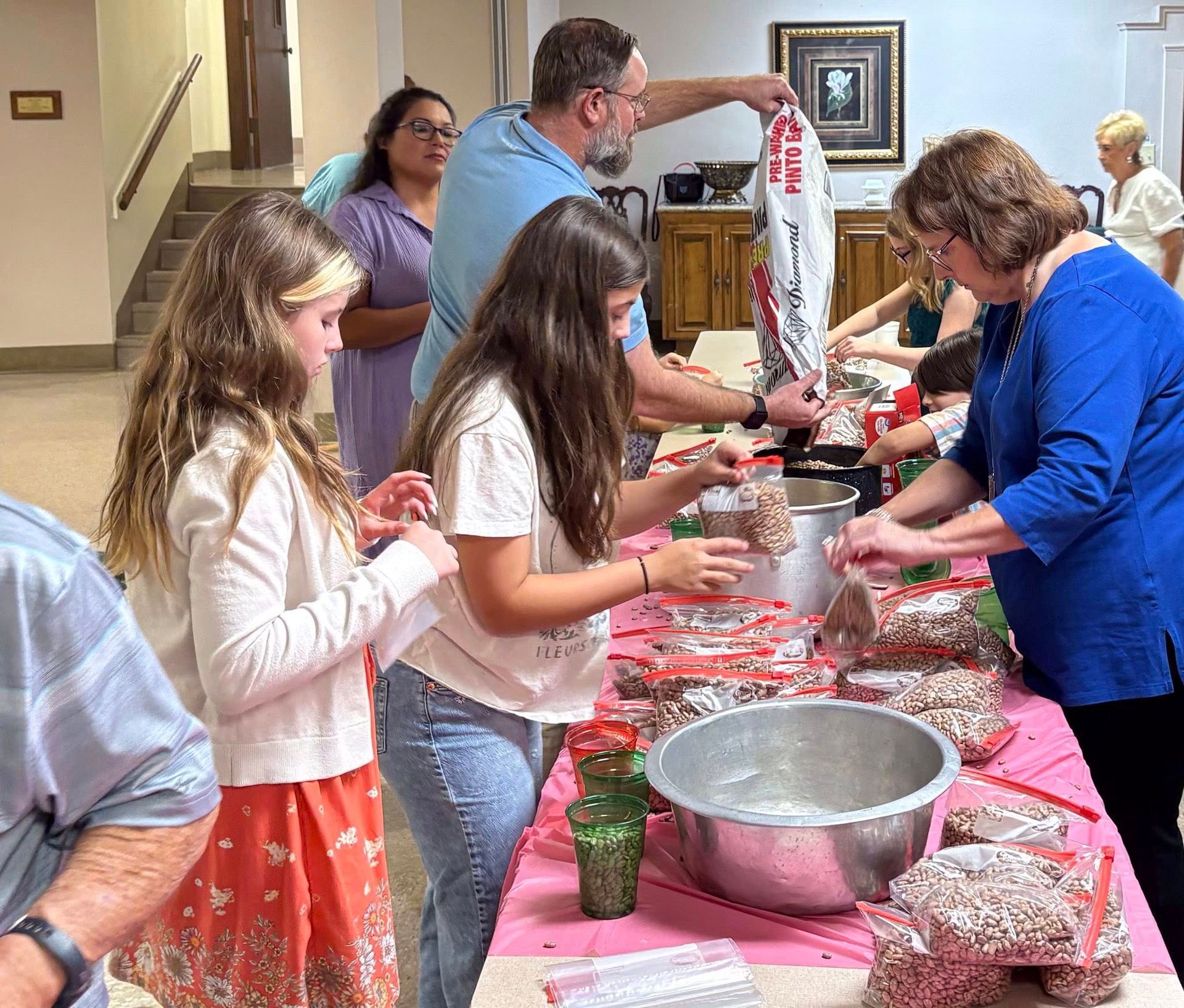 People filling bags with items at a table. Indoor setting, with a staircase visible in the background.