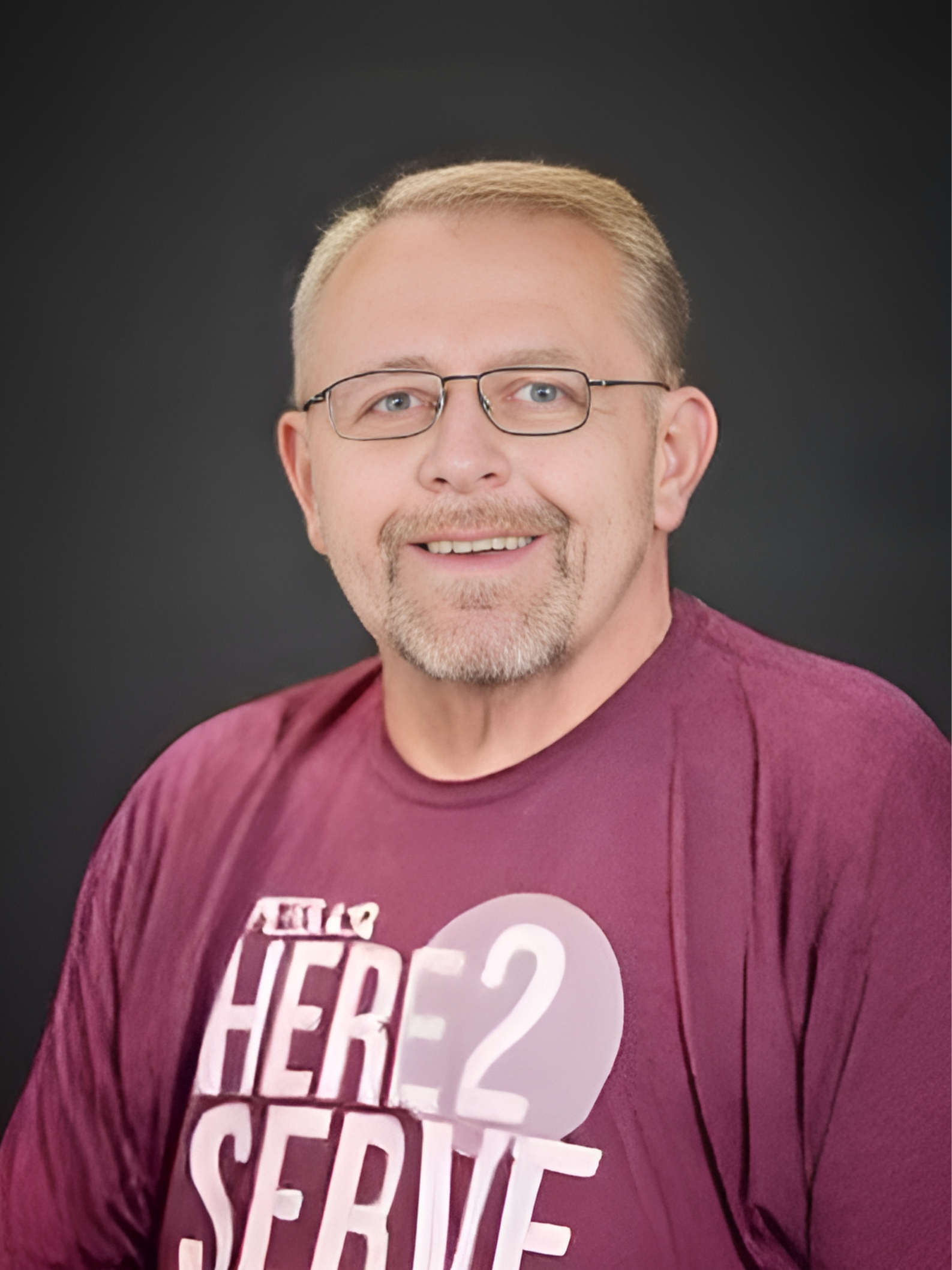 A man with glasses and a beard is wearing a purple shirt that says `` here 2 serve ''.