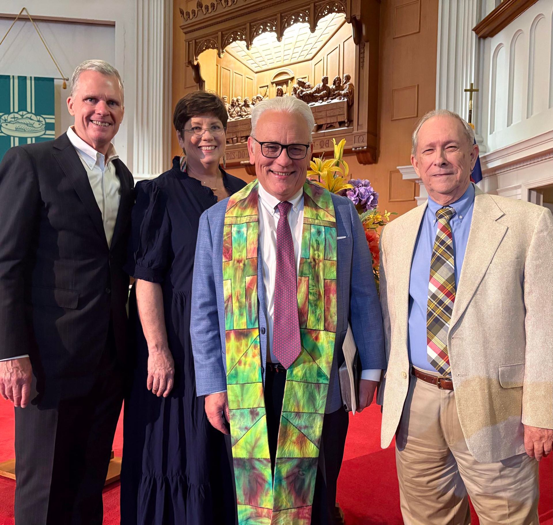 Four people pose in a church. A man in a blue jacket and colorful stole stands with three others.