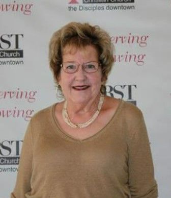 A woman wearing glasses and a necklace is standing in front of a wall that says serving the disciples downtown.
