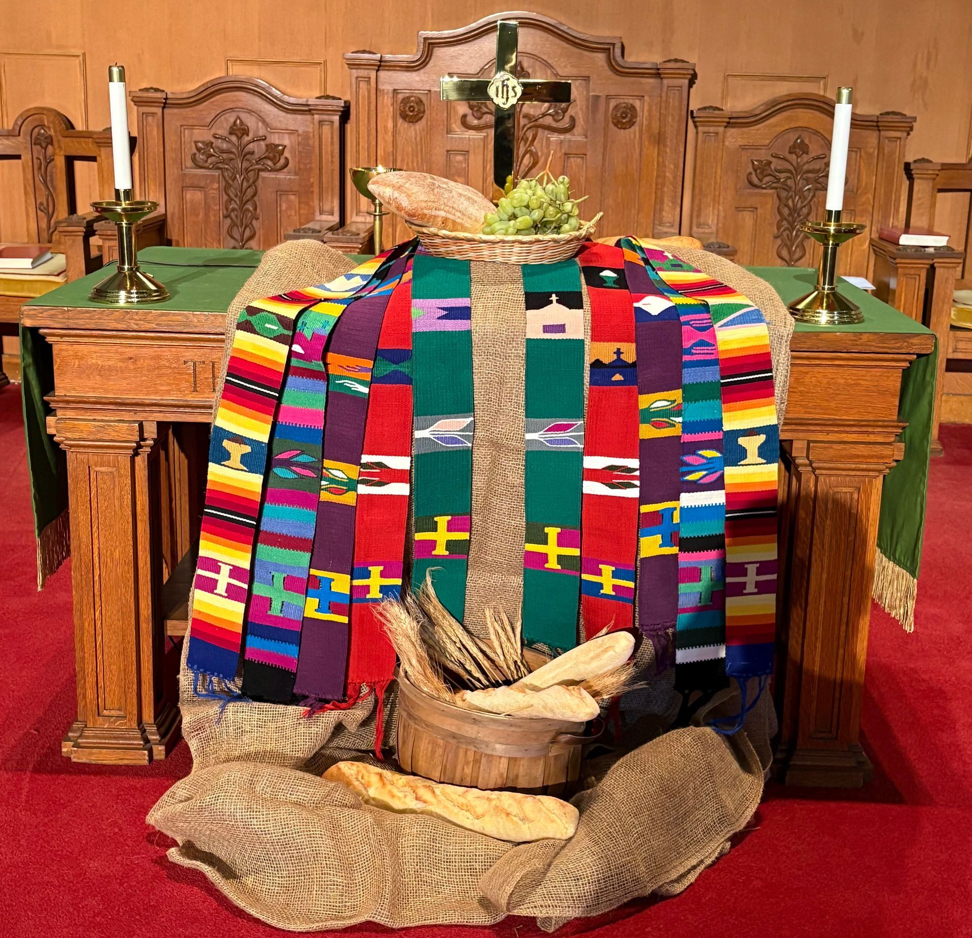 Altar with burlap, colorful cloth, bread, wheat, and candles. Church setting with wooden backdrop and a cross.