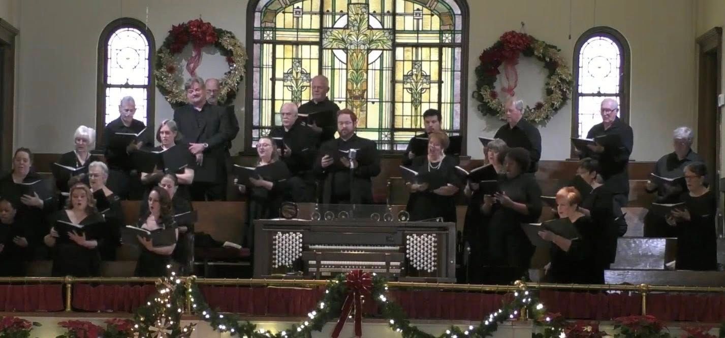 Choir singing in a church, decorated for Christmas. Group wears black clothing. Large stained-glass window behind them.