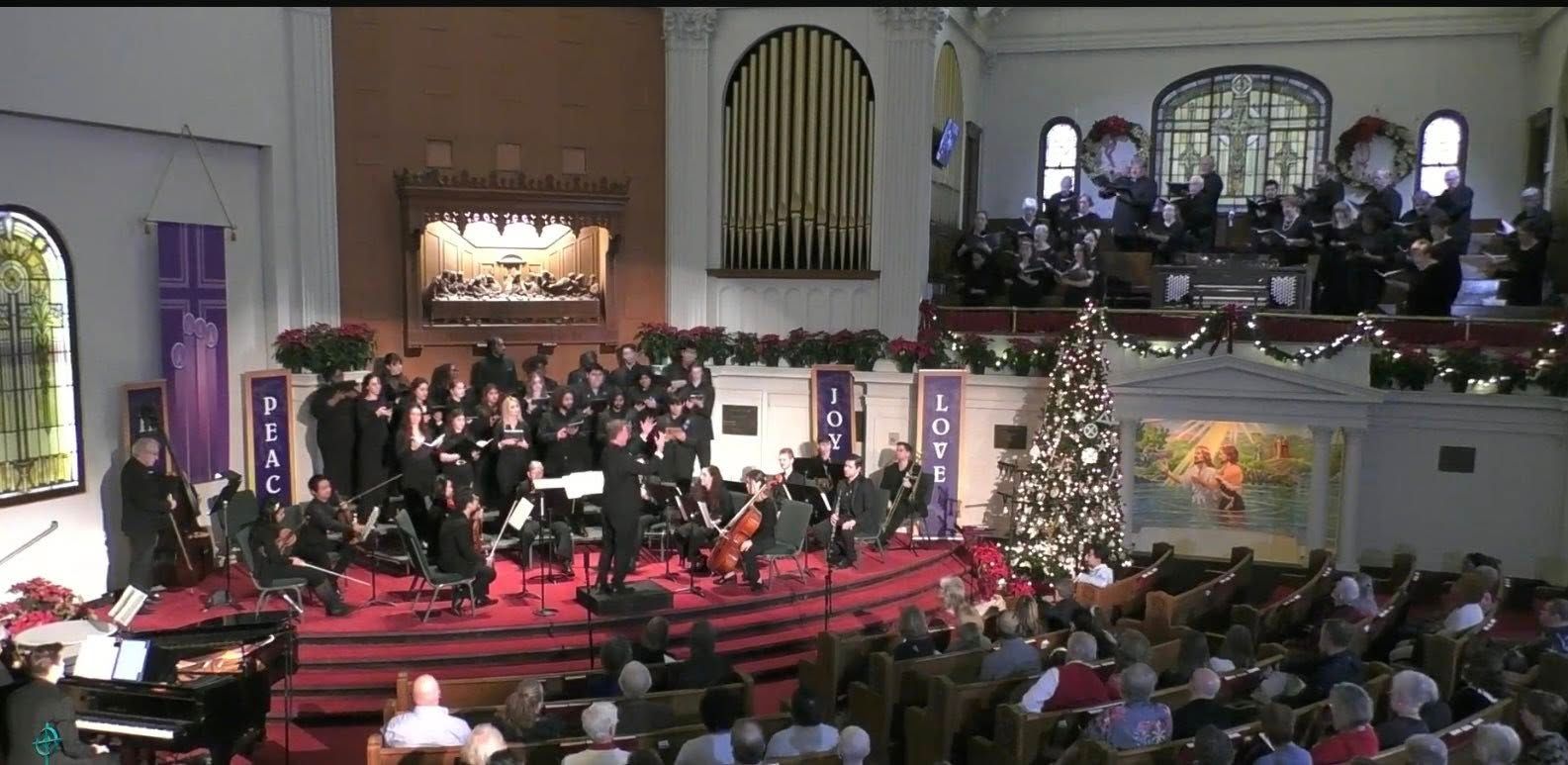 A choir and orchestra perform on a stage in a church during a Christmas concert, decorated with a Christmas tree.
