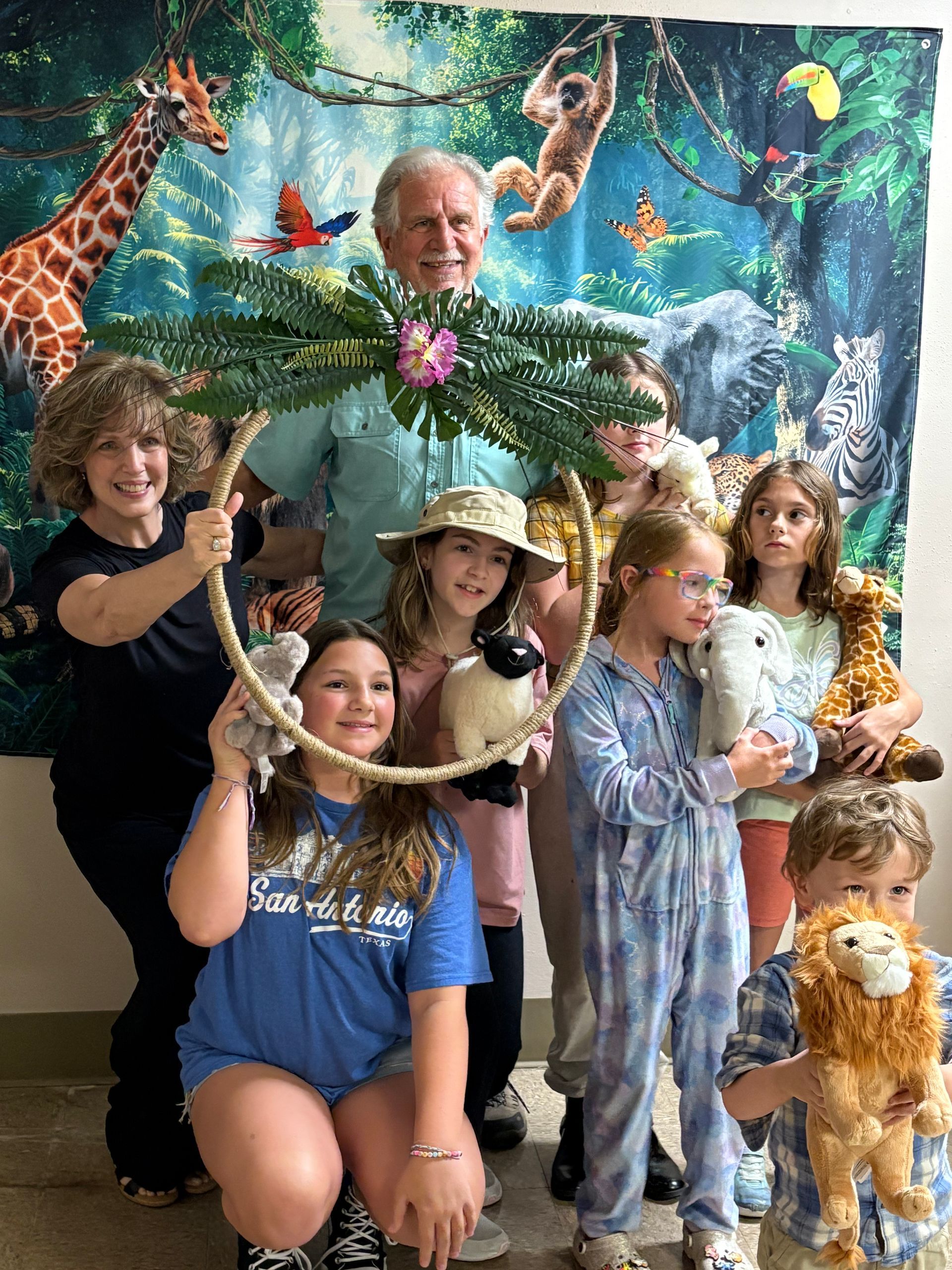 Group poses with stuffed animals, jungle backdrop. Smiling adults and children hold a decorated hoop.