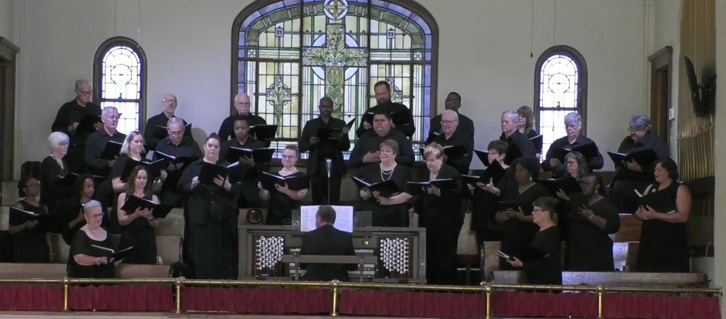 Choir performing in a church, facing forward. They are on a raised platform, singing from sheet music.