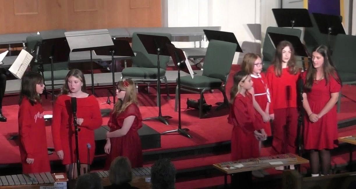 Choir of girls in red dresses on a stage with music stands, performing in a church.