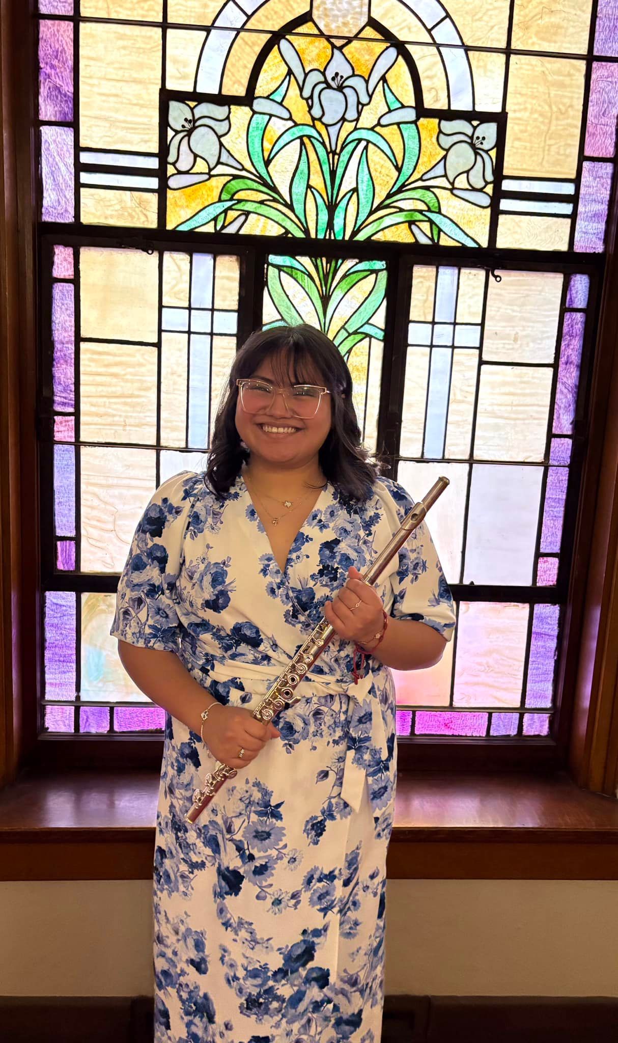 Woman in blue floral dress holding a flute, smiling in front of a stained-glass window.