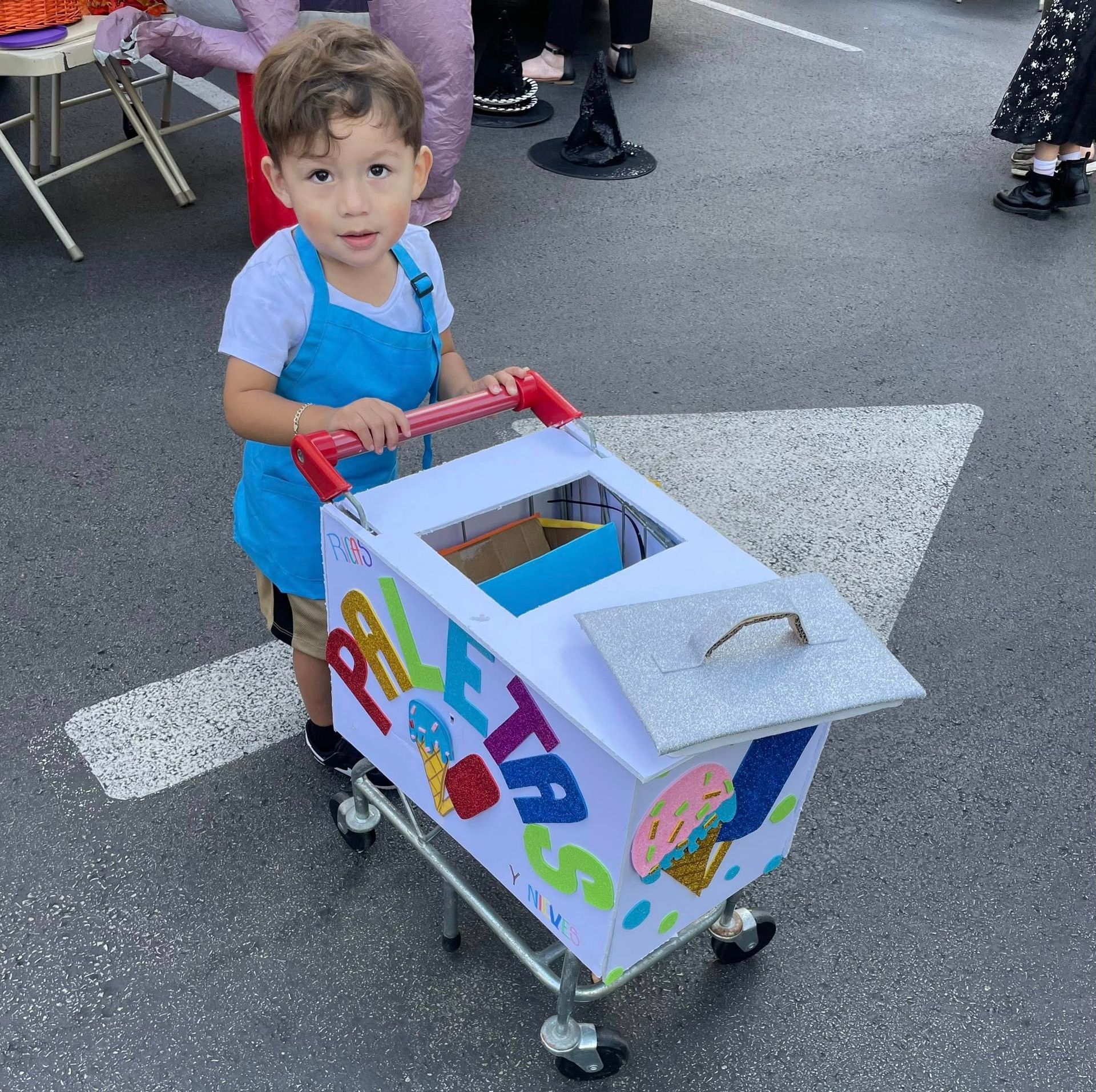 Child in blue apron costume pushes a cart decorated to look like an ice cream stand.