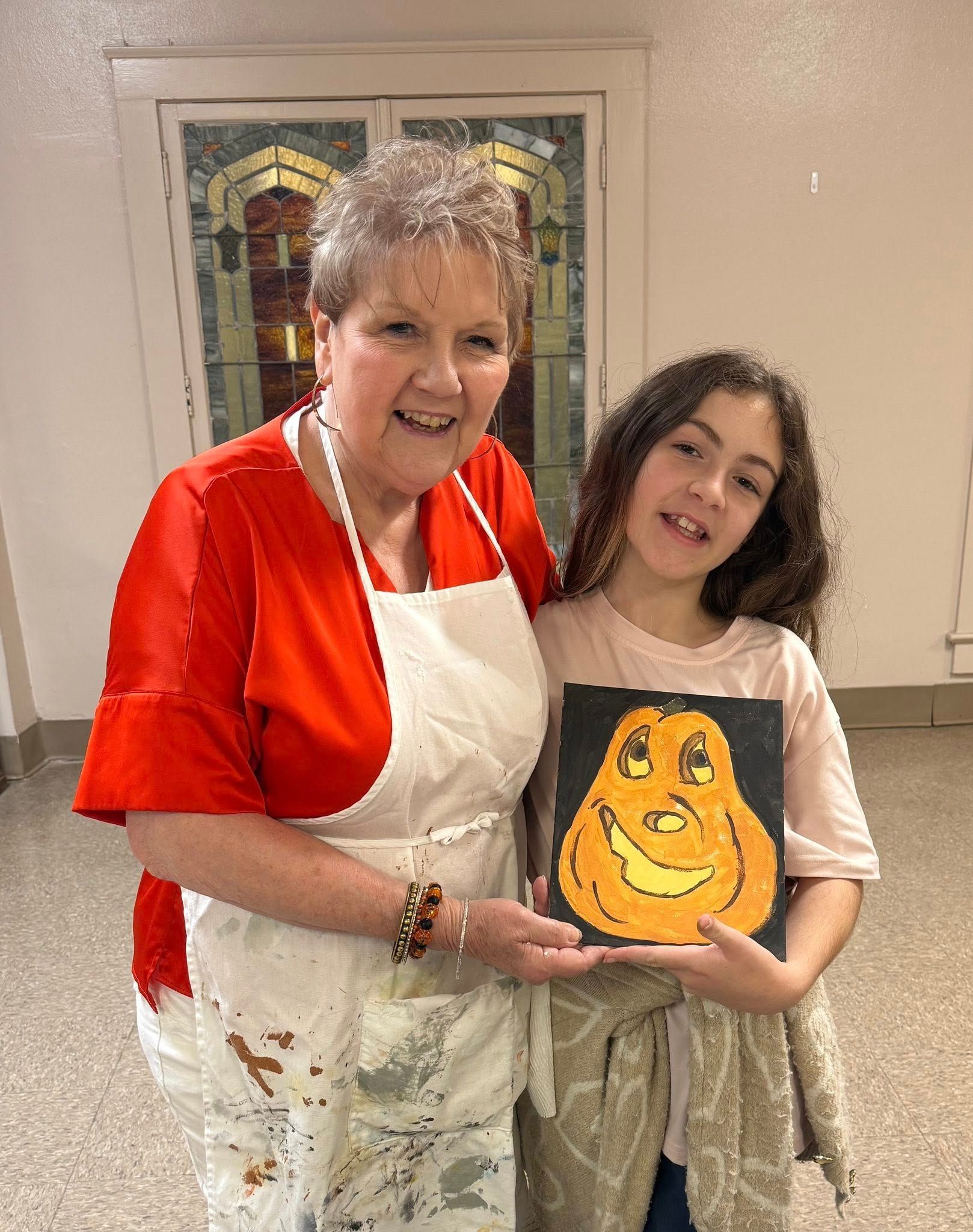 Woman and girl holding a painted pumpkin. Woman wears red top and apron. Girl smiles, holding the artwork.