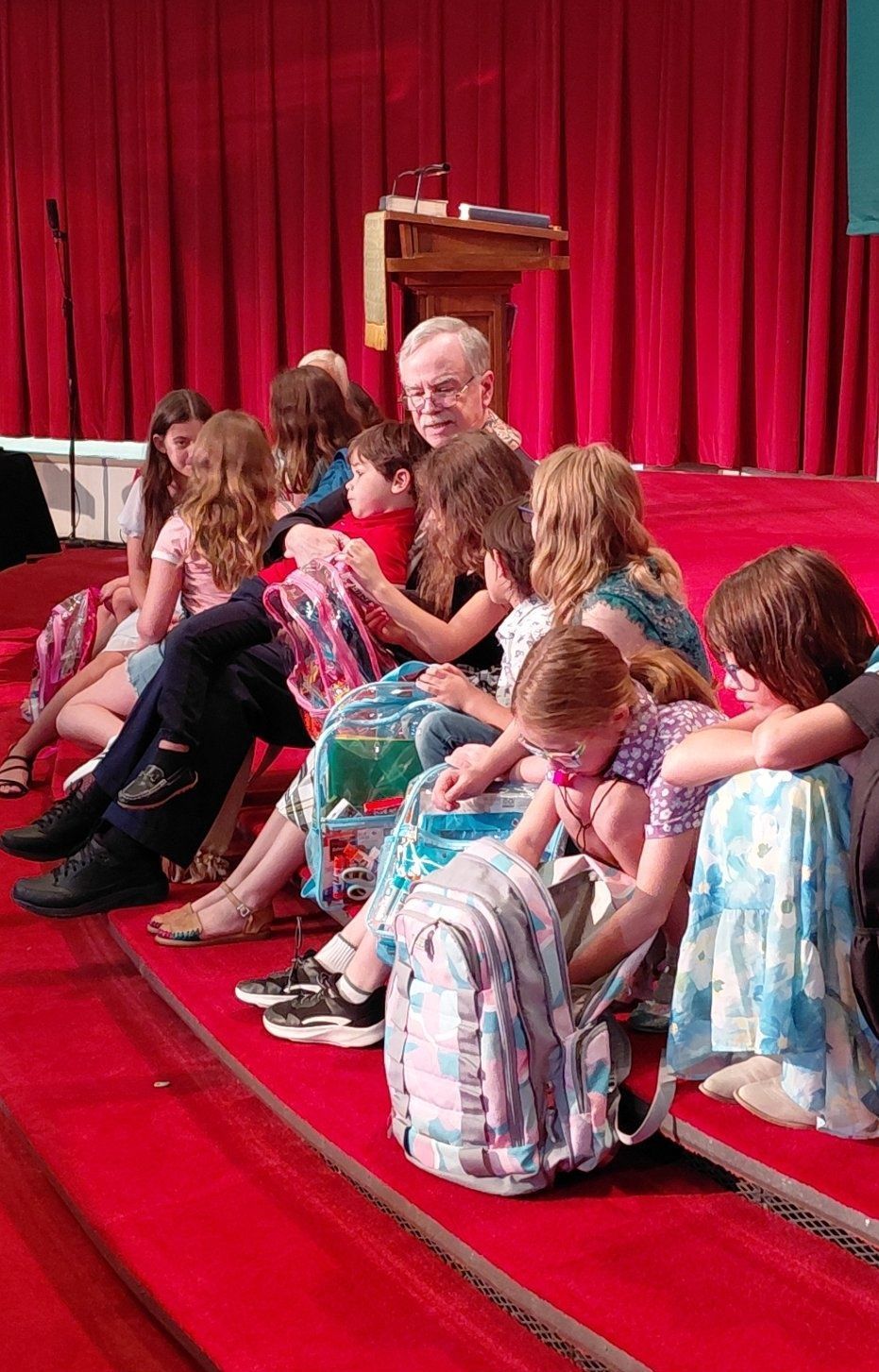 Group of people sitting on red steps, some with backpacks, near a stage with red curtains.
