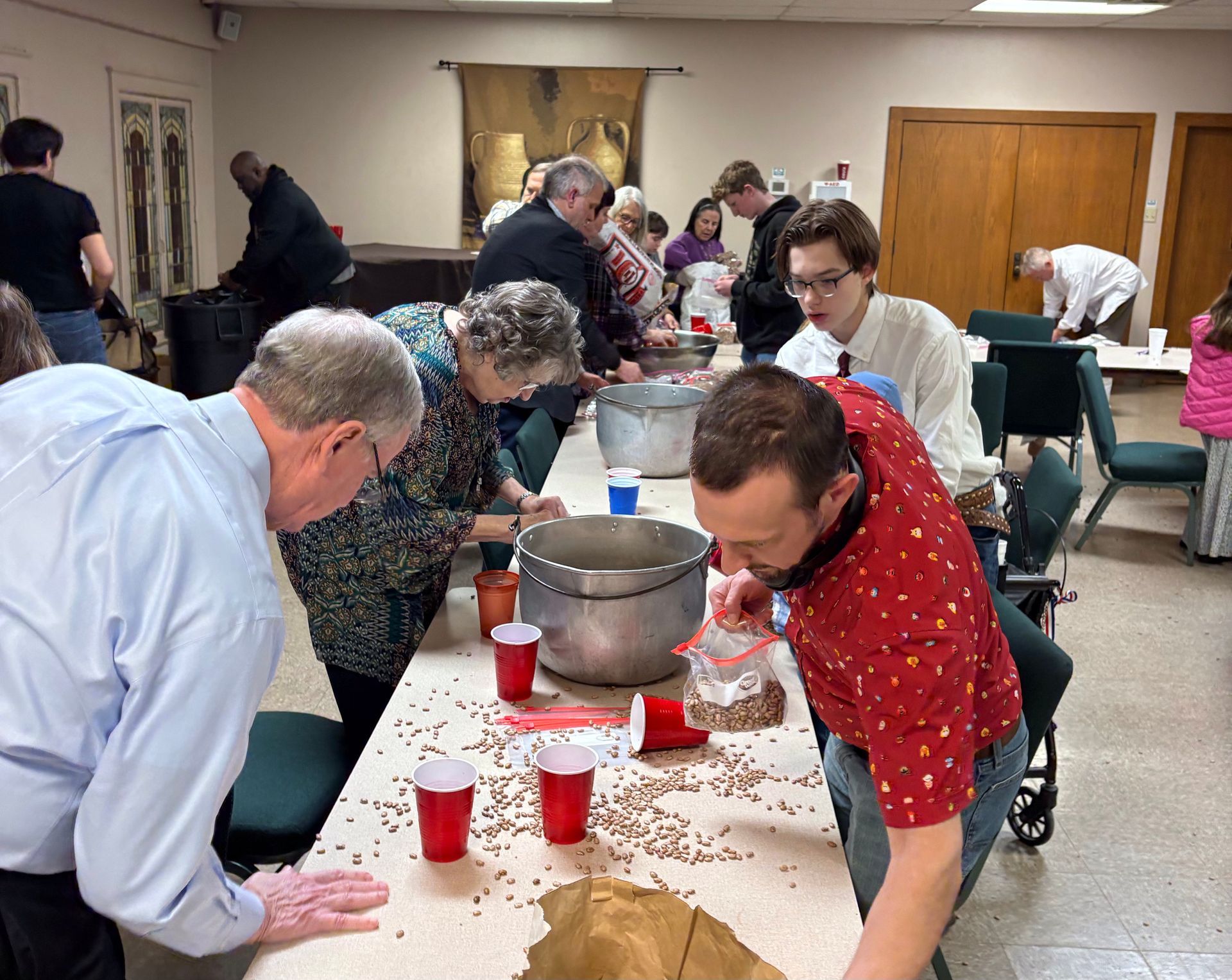 People sorting beans at a table; red cups, pot, and bags are present in a church-like setting.