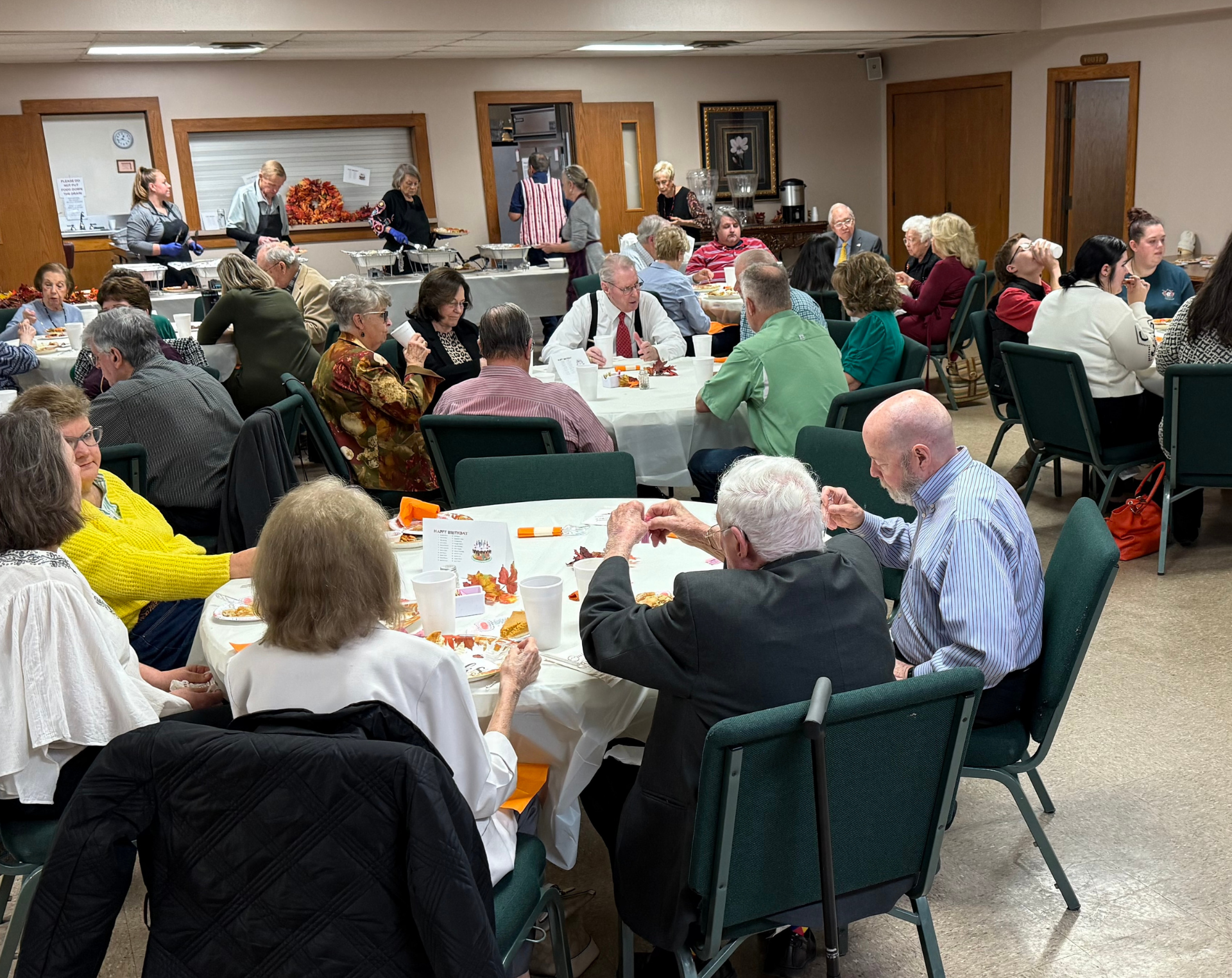 People seated at round tables in a community center, likely at a social gathering or meal.
