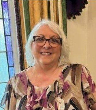 A woman wearing glasses and a floral shirt is smiling in front of a stained glass window.