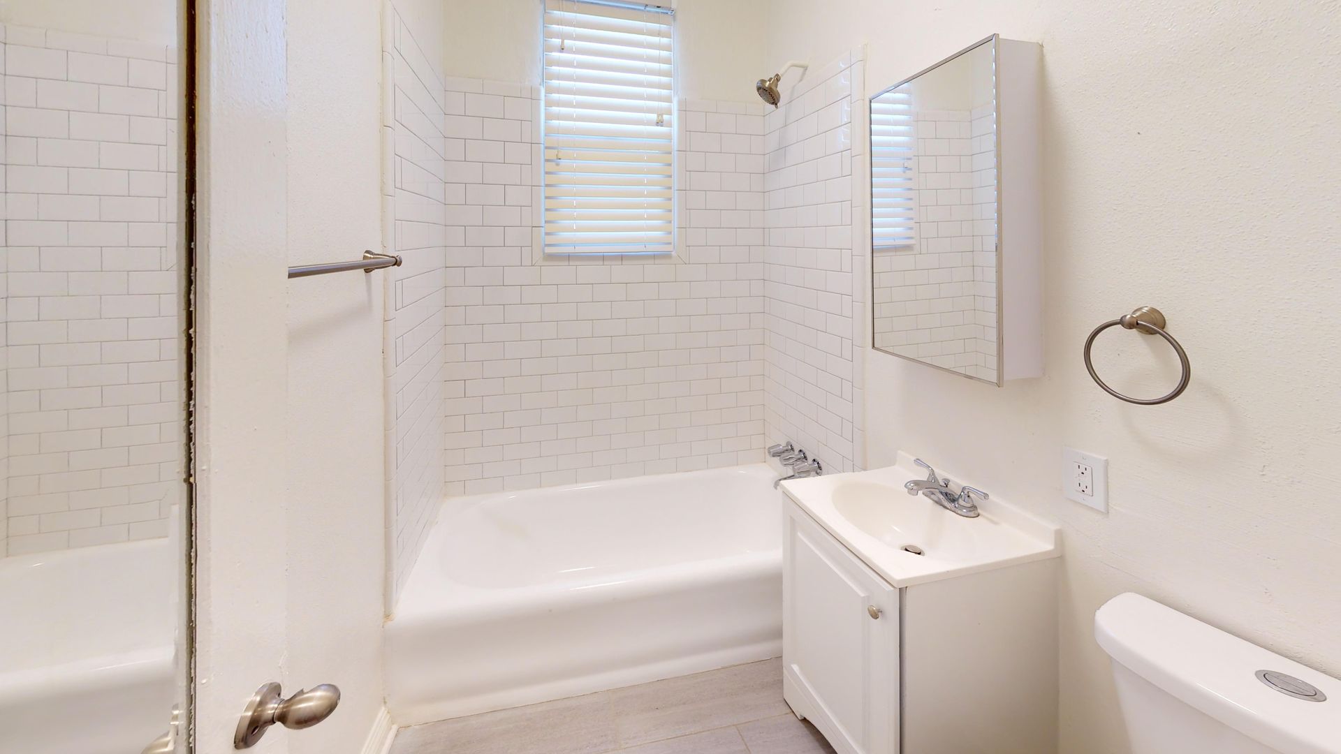 Bathroom with white walls, tub, sink, and toilet. A window with blinds is centered above the tub.