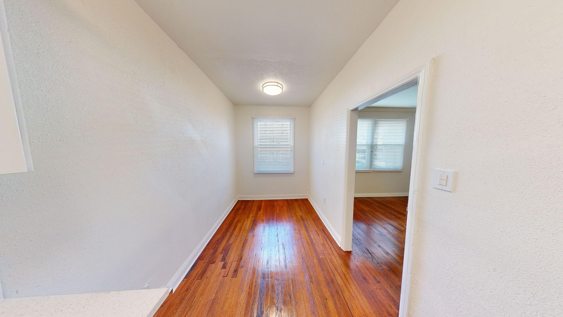 Hallway with hardwood floors, white walls, and a window at the end. An open doorway on the right.