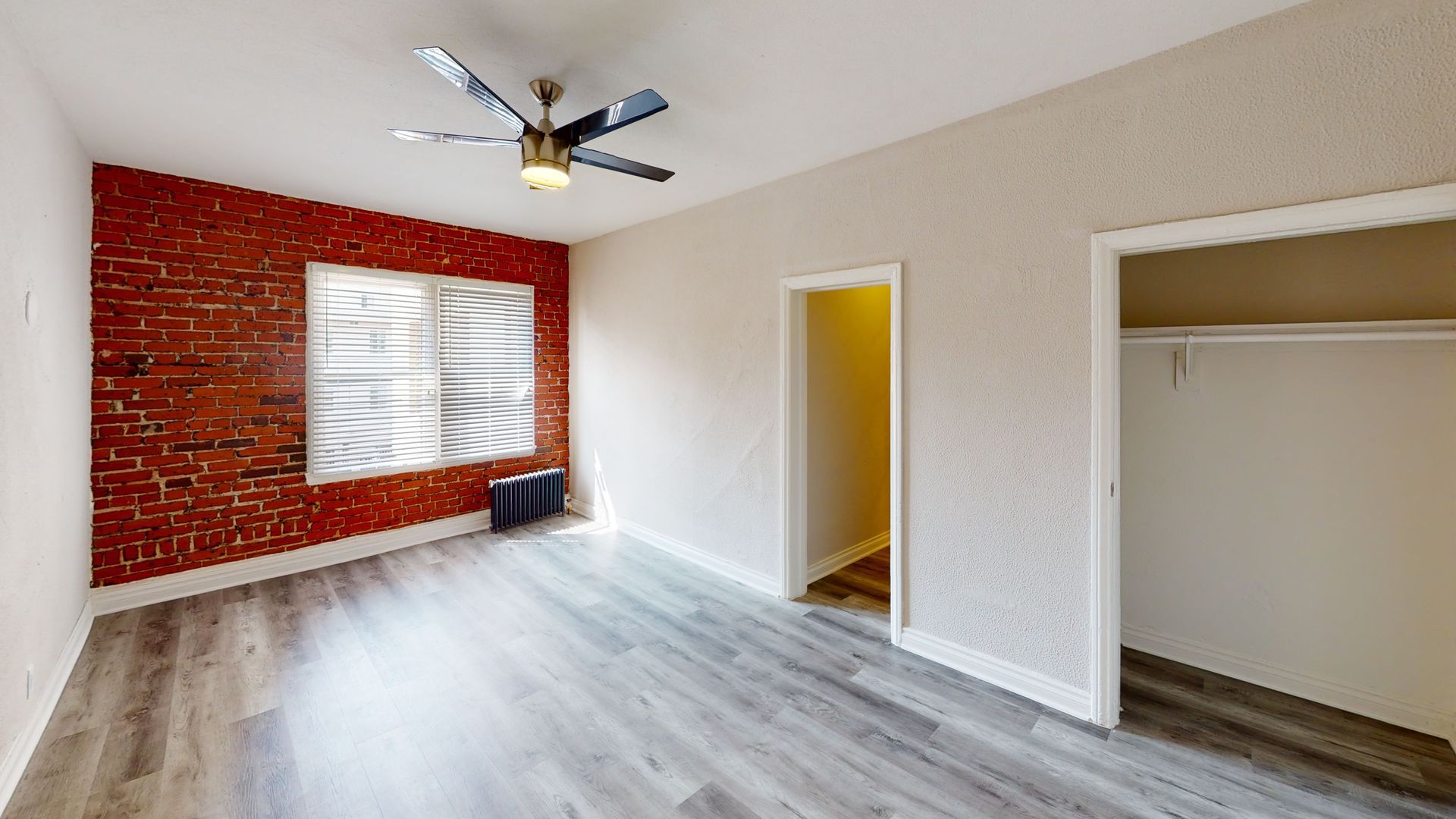 Empty room with exposed brick wall, window, gray flooring, open closet, and ceiling fan.